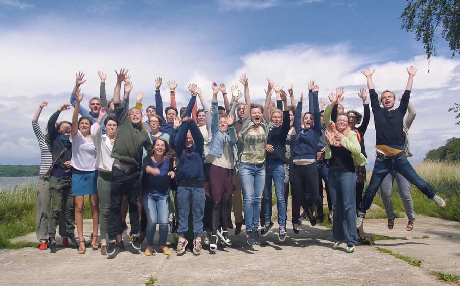Teamende des teamGLOBAL springen für ein Gruppenfoto in gemeinsam die Luft. Standbild eines Imagefilms von Brandenburg Film Foto.
